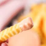 A crinkle-cut fry, dipped in a rich fry sauce, is held up close, with a plate of fries blurred in the background.