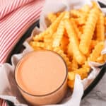 Crinkle-cut fries in a basket paired with a glass cup of pink fry sauce, all set on a white marble surface. A red and white striped cloth adds charm in the background.