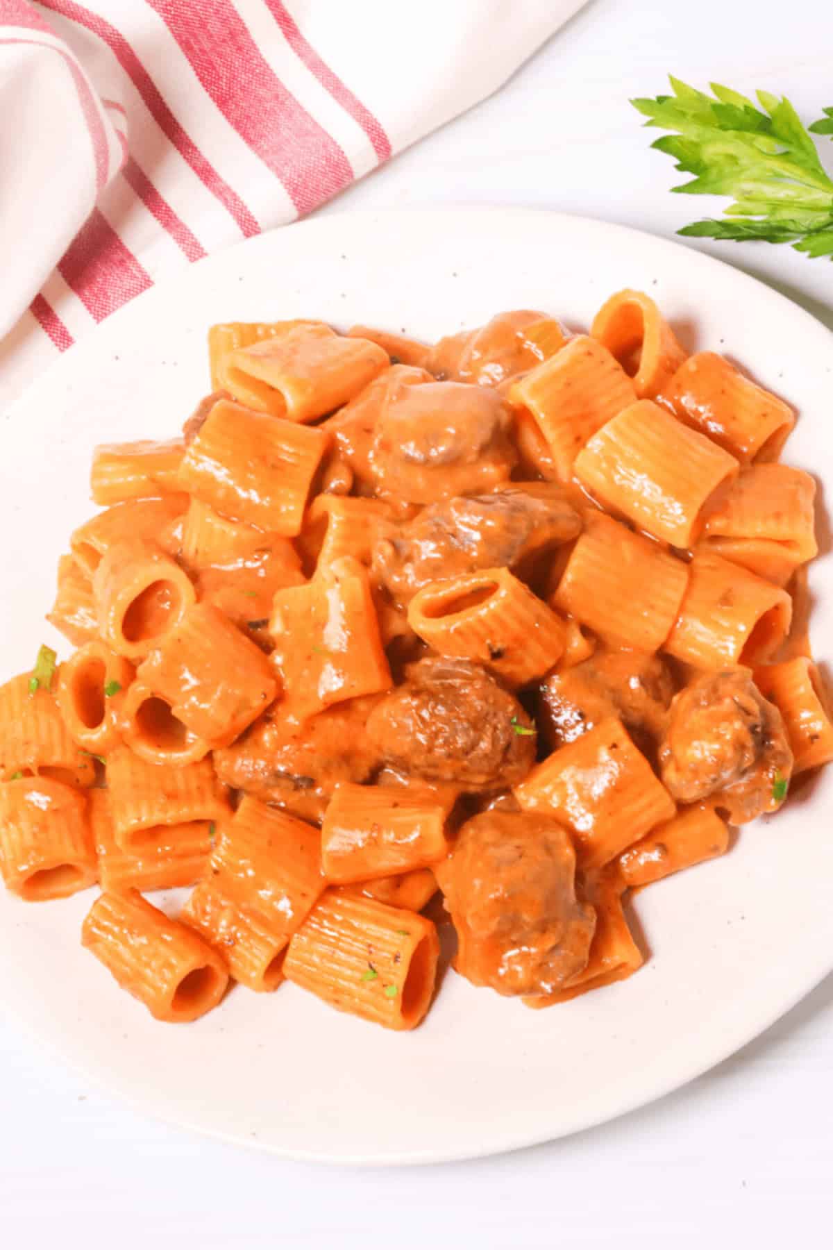 A plate of Italian pasta with a tomato-based sauce, meatballs, and savory pork sausage is garnished with a sprig of parsley.