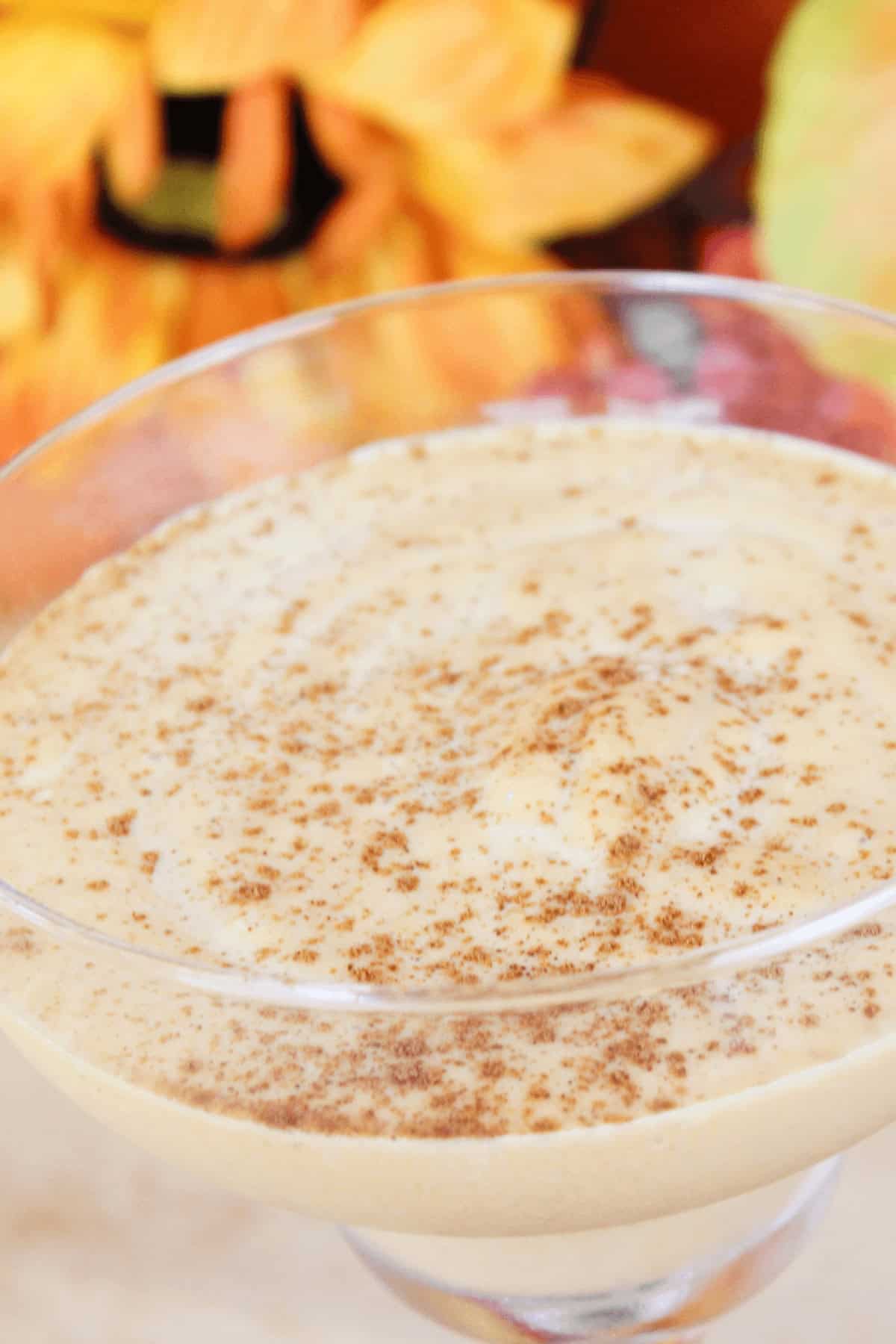 A Pumpkin Spice White Russian, this creamy dessert in a glass is topped with a sprinkle of cinnamon, perfectly complementing the blurred autumn-themed decorations in the background.