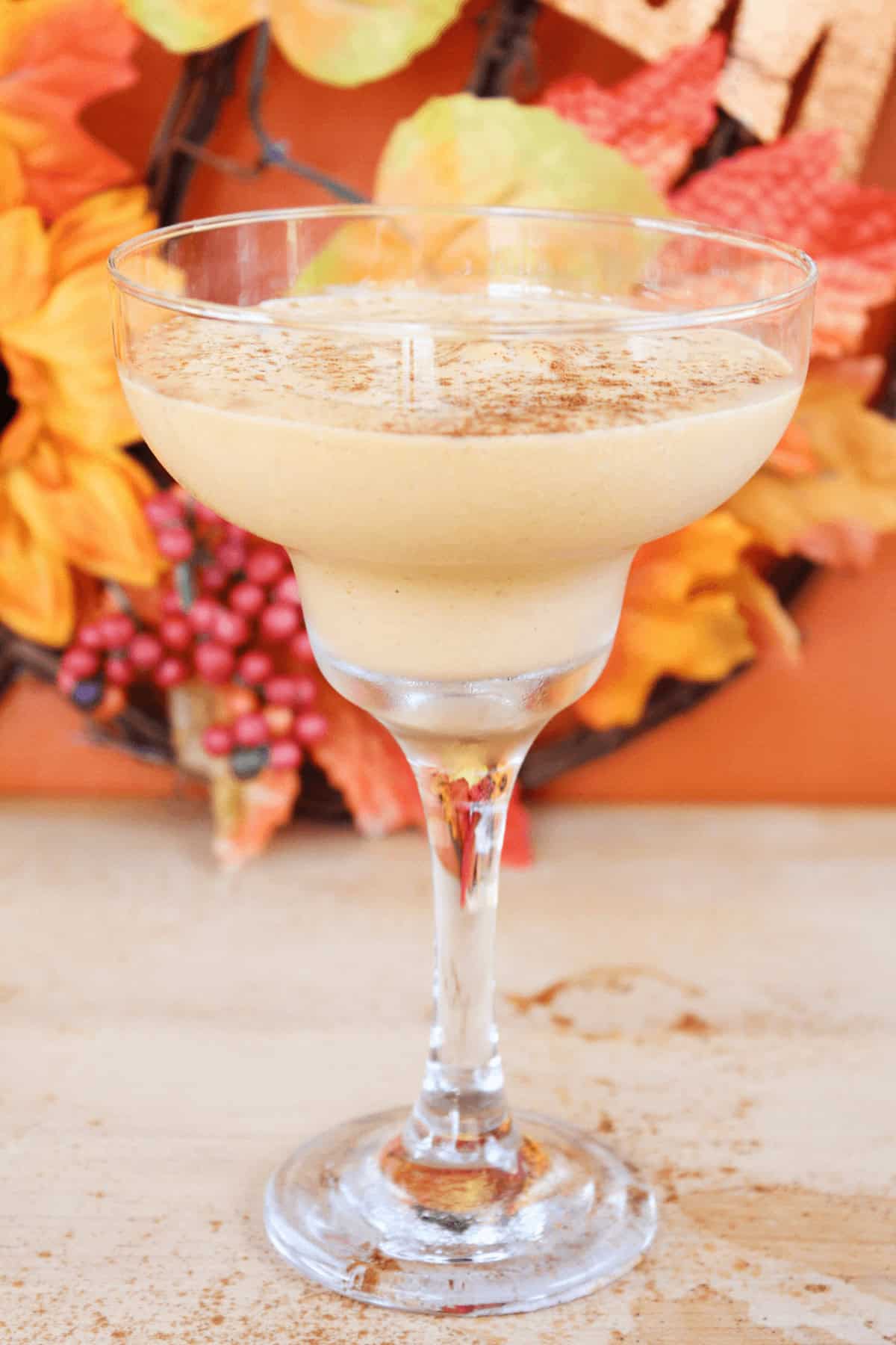 A creamy Pumpkin Spice White Russian in a margarita glass, topped with a sprinkle of cinnamon, set against an autumn-themed background with orange leaves and berries.