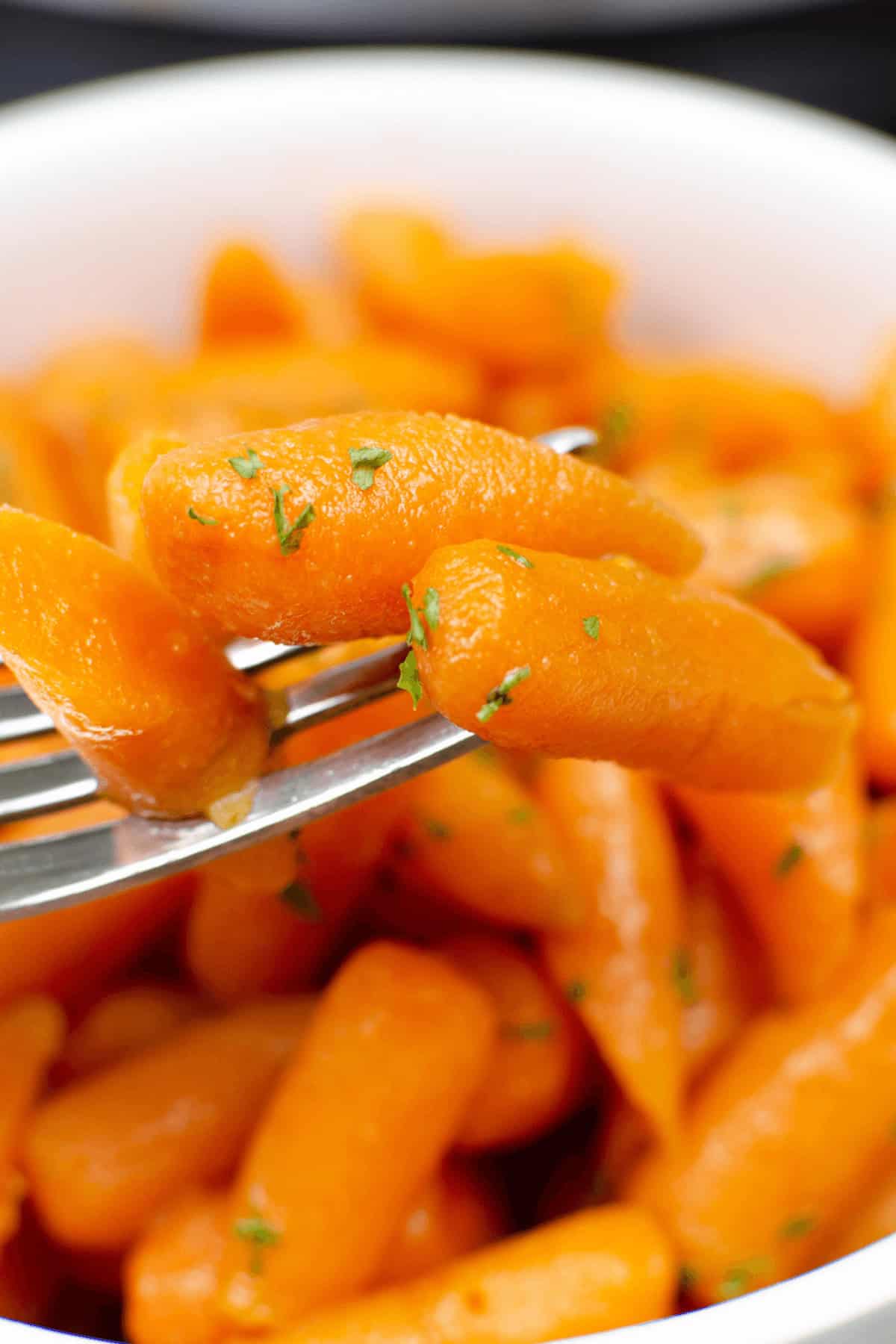 A close-up of a fork lifting a glazed baby carrot from a bowl filled with more Instant Pot carrots, garnished with chopped herbs.
