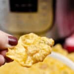 A hand holding a tortilla chip doused in Crock Pot Queso Dip hovers enticingly in front of a blurred background, possibly suggesting the presence of a kitchen appliance.
