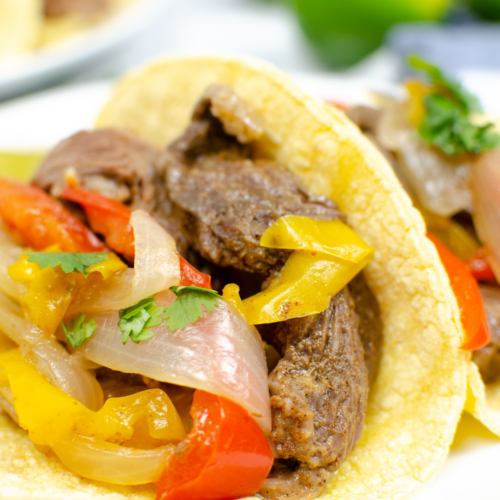 Close-up of an Instant Pot beef fajita taco with colorful bell peppers and onions in a soft corn tortilla, garnished with cilantro, on a white plate.