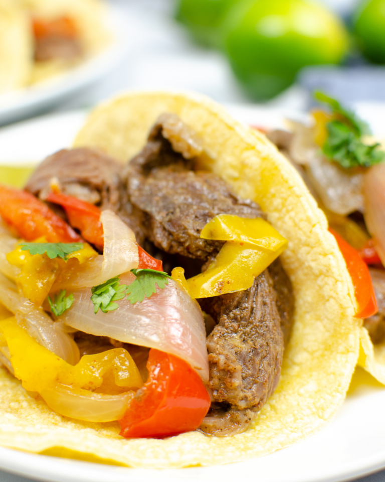 Close-up of an Instant Pot beef fajita taco with colorful bell peppers and onions in a soft corn tortilla, garnished with cilantro, on a white plate.