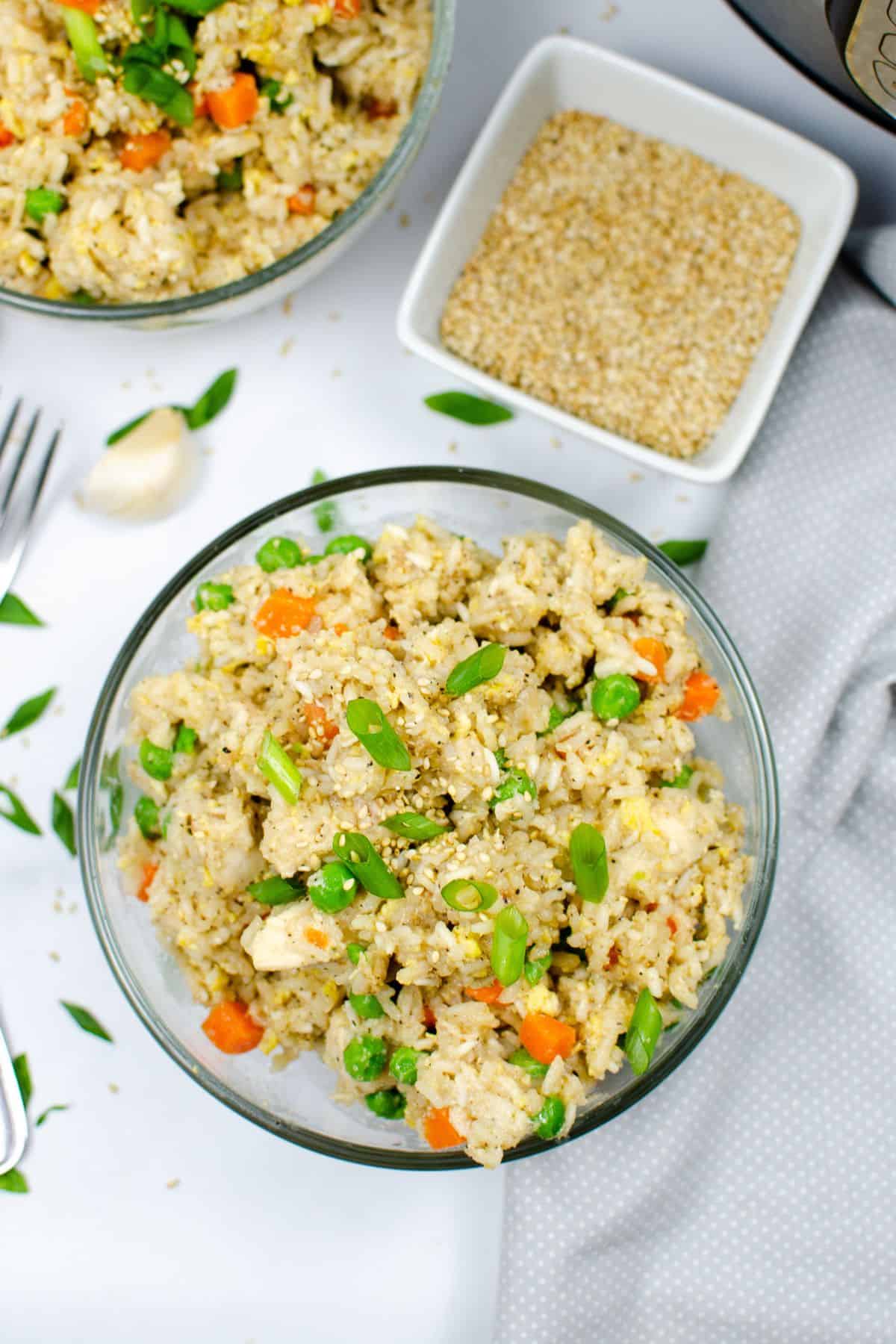 A bowl of Instant Pot chicken fried rice with peas, carrots, scrambled egg, and green onions, served beside a small dish of sesame seeds on a white surface.