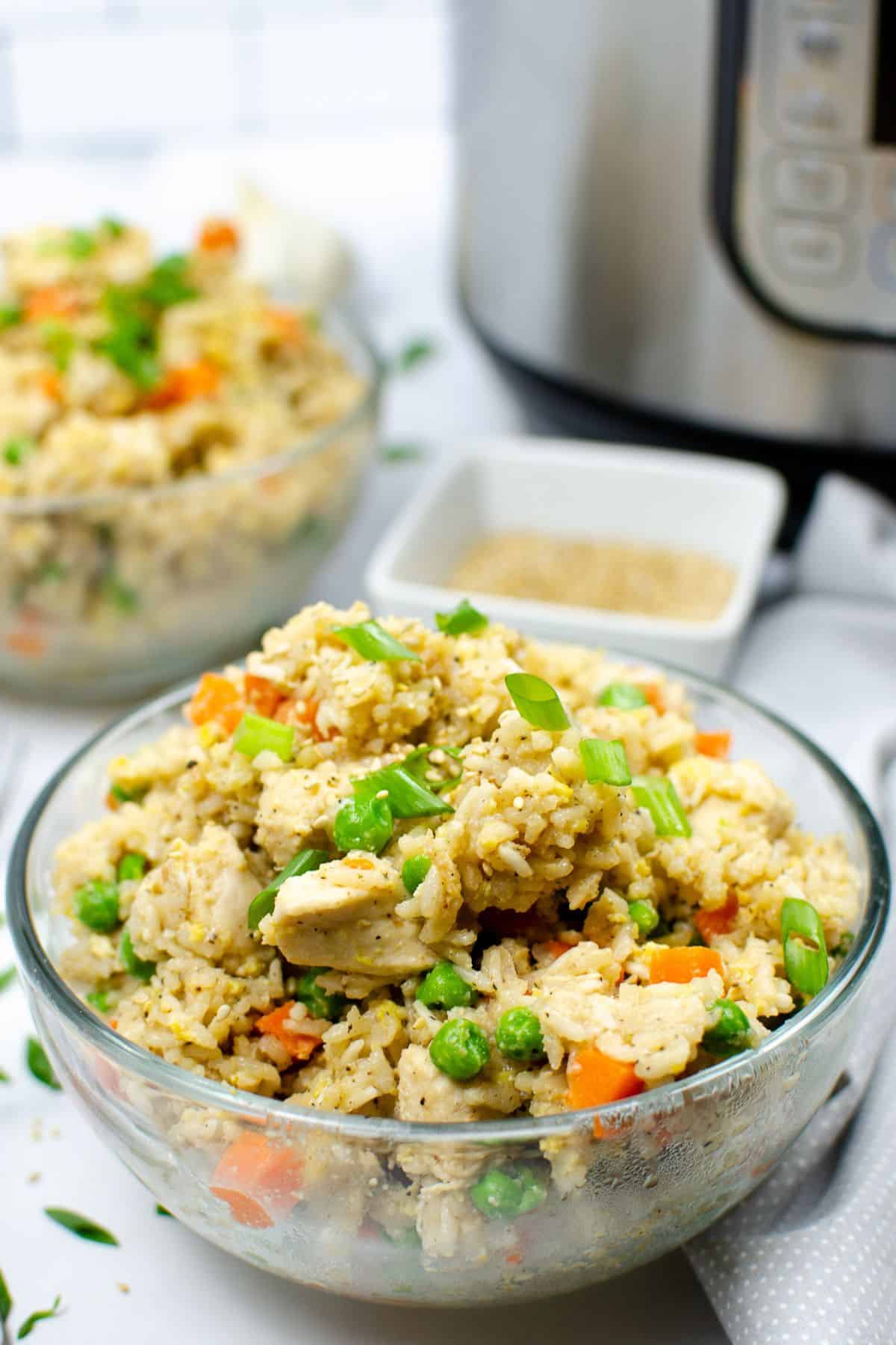 A glass bowl filled with instant pot chicken fried rice featuring peas, carrots, and green onions, with a second bowl and an Instant Pot in the background.