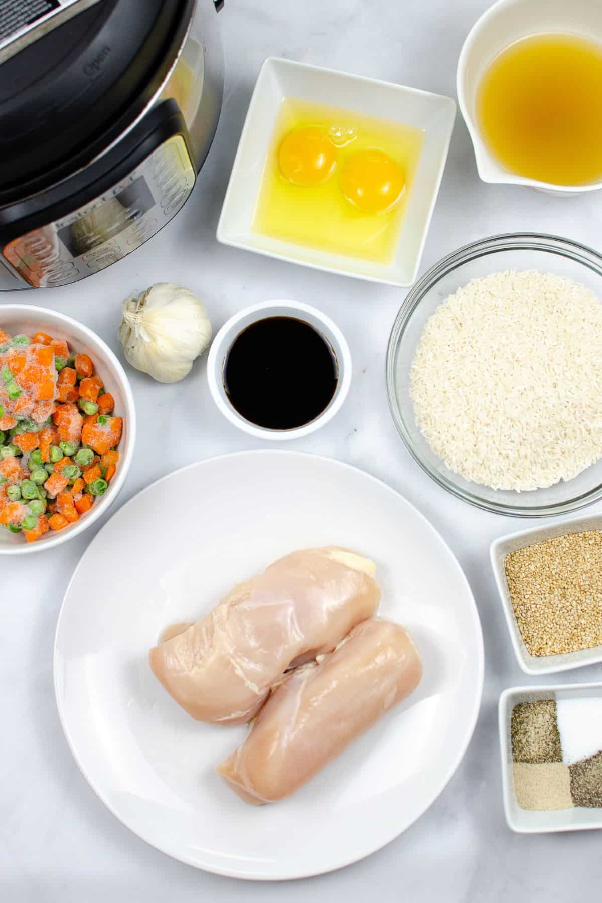 Top-down view of ingredients —raw chicken breasts, eggs, rice, frozen mixed vegetables, garlic, broth, soy sauce, and assorted seasonings—arranged on a white surface next to a pressure cooker.