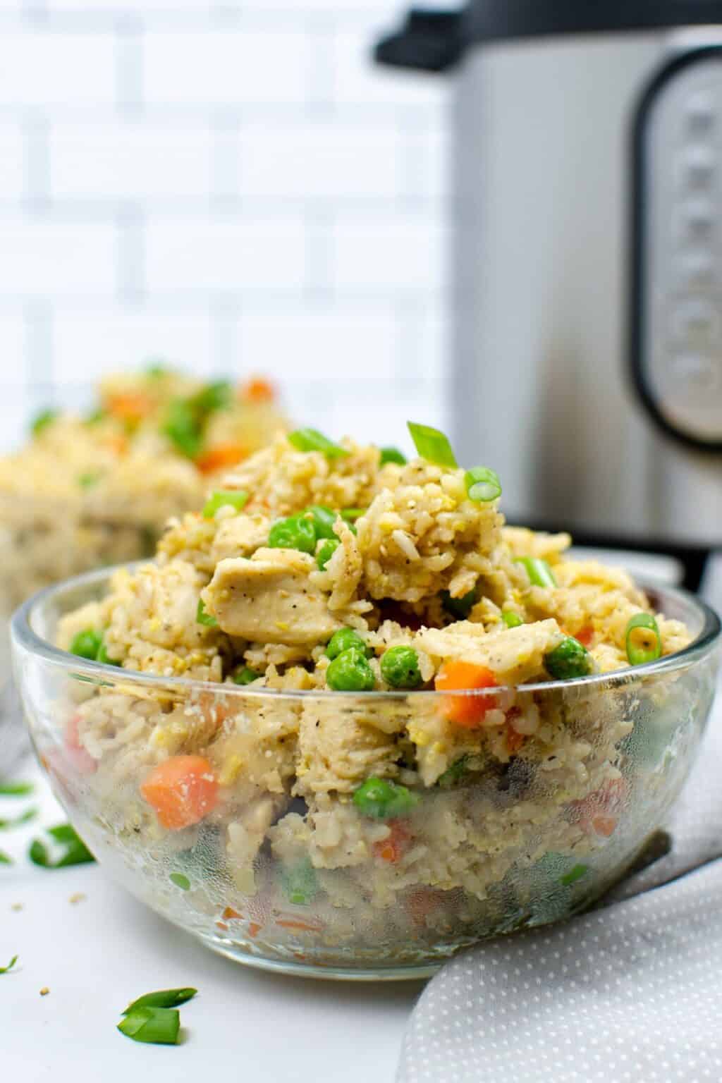 A clear glass bowl filled with Instant Pot chicken fried rice, featuring peas, carrots, and green onions, sits in the foreground with a kitchen appliance in the background.