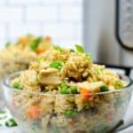 A clear glass bowl filled with Instant Pot chicken fried rice, featuring peas, carrots, and green onions, sits in the foreground with a kitchen appliance in the background.