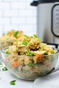 A clear glass bowl filled with Instant Pot chicken fried rice, featuring peas, carrots, and green onions, sits in the foreground with a kitchen appliance in the background.