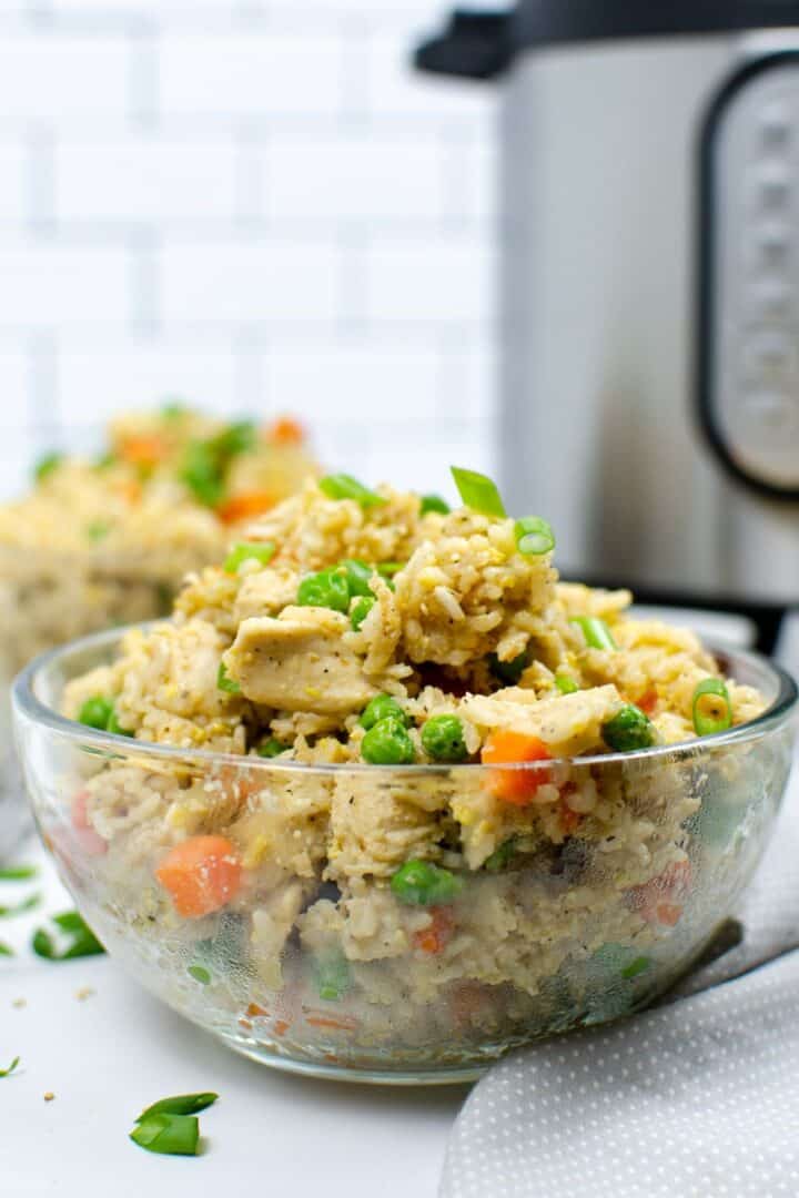 A clear glass bowl filled with Instant Pot chicken fried rice, featuring peas, carrots, and green onions, sits in the foreground with a kitchen appliance in the background.
