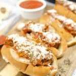 Enjoy a delectable meatball subs recipe featuring juicy meatballs smothered in rich tomato sauce and topped with grated cheese. Presented elegantly on a wooden board, it's accompanied by a tempting bowl of extra sauce in the background.