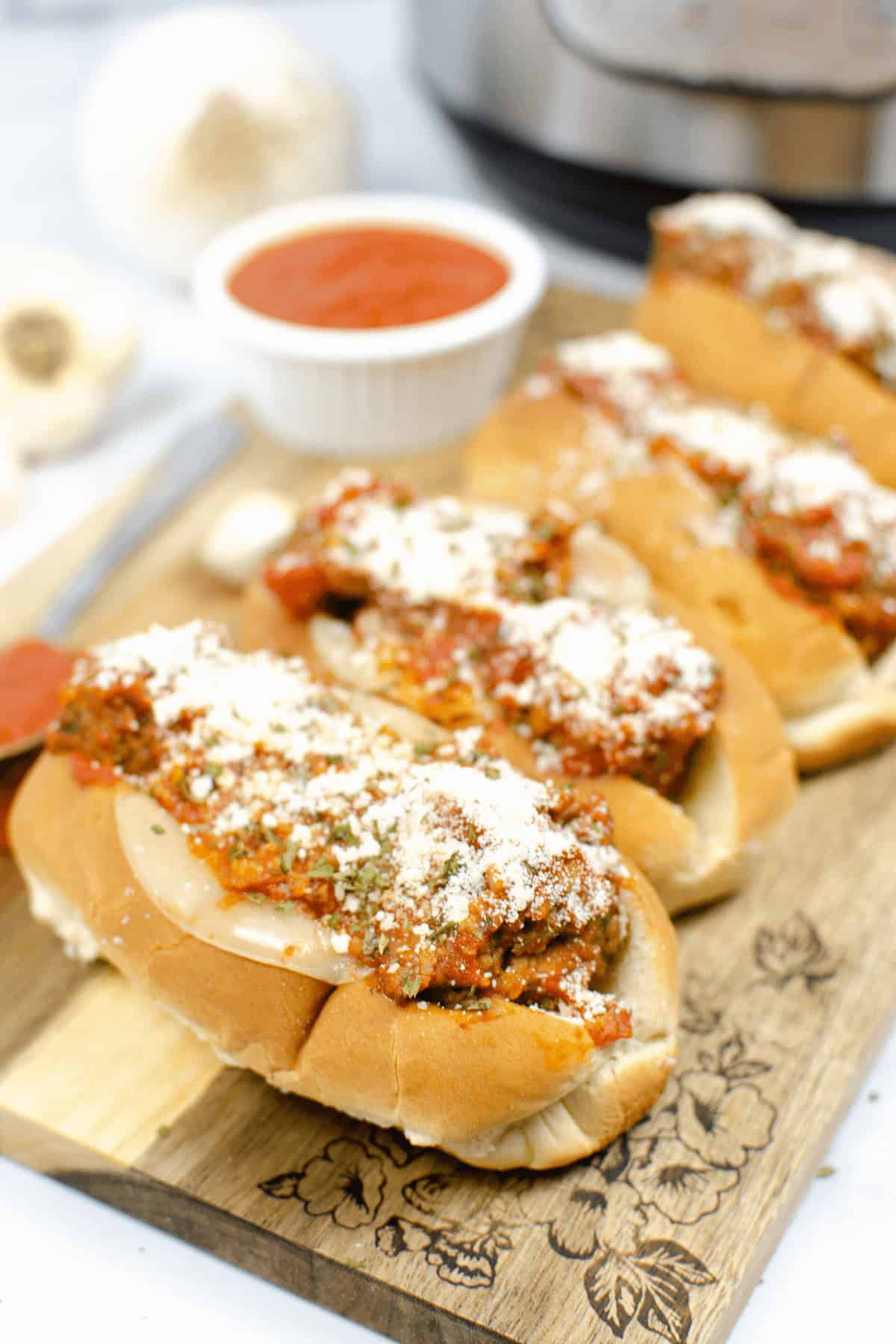 Enjoy a delectable meatball subs recipe featuring juicy meatballs smothered in rich tomato sauce and topped with grated cheese. Presented elegantly on a wooden board, it's accompanied by a tempting bowl of extra sauce in the background.