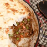 A red baking dish with shepherd's pie showcases a delicious masterpiece, featuring mashed potato topping over a savory filling of ground beef, peas, and carrots. A plaid cloth sits invitingly beside this tempting shepherd's pie recipe.