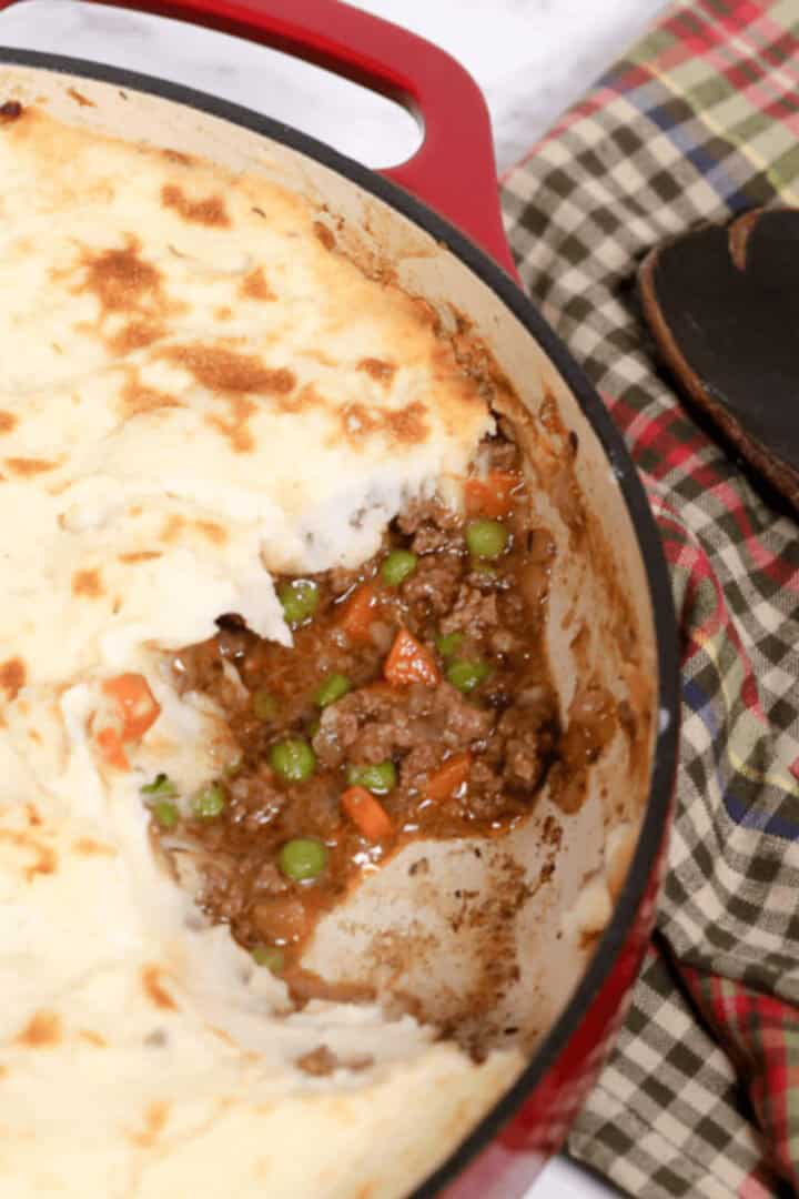 A red baking dish with shepherd's pie showcases a delicious masterpiece, featuring mashed potato topping over a savory filling of ground beef, peas, and carrots. A plaid cloth sits invitingly beside this tempting shepherd's pie recipe.