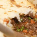 Close-up of a shepherd's pie in a red dish, showcasing a scooped portion to reveal the delicious layers in this classic recipe with ground beef, peas, and carrots beneath a perfectly golden layer of mashed potatoes.