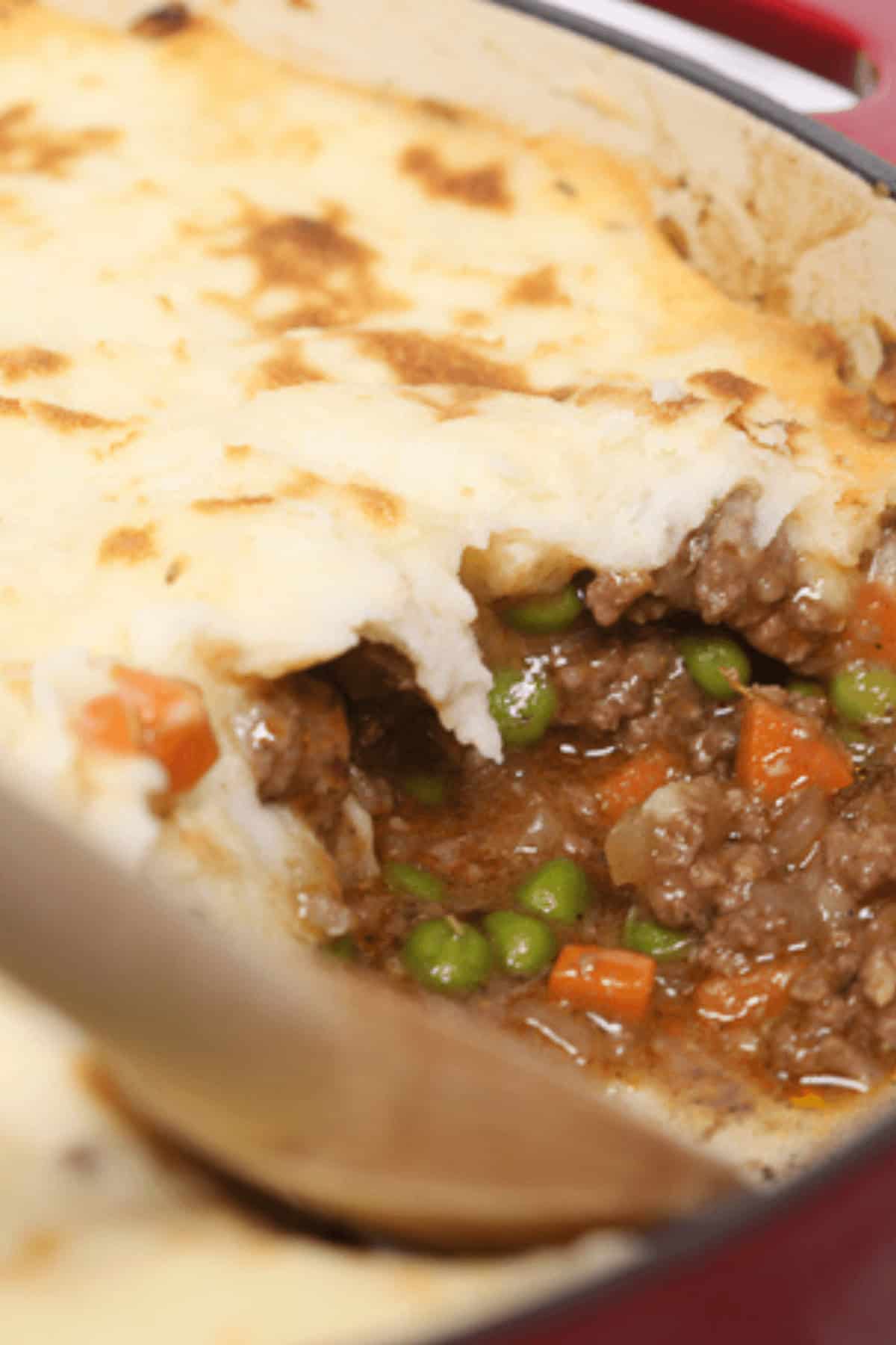 Close-up of a shepherd's pie in a red dish, showcasing a scooped portion to reveal the delicious layers in this classic recipe with ground beef, peas, and carrots beneath a perfectly golden layer of mashed potatoes.