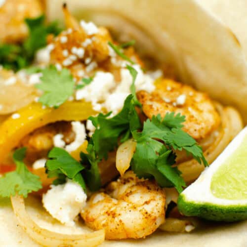 A shrimp fajita taco garnished with cilantro, crumbled cheese, sautéed onions and yellow peppers, served with a lime wedge on a white plate.