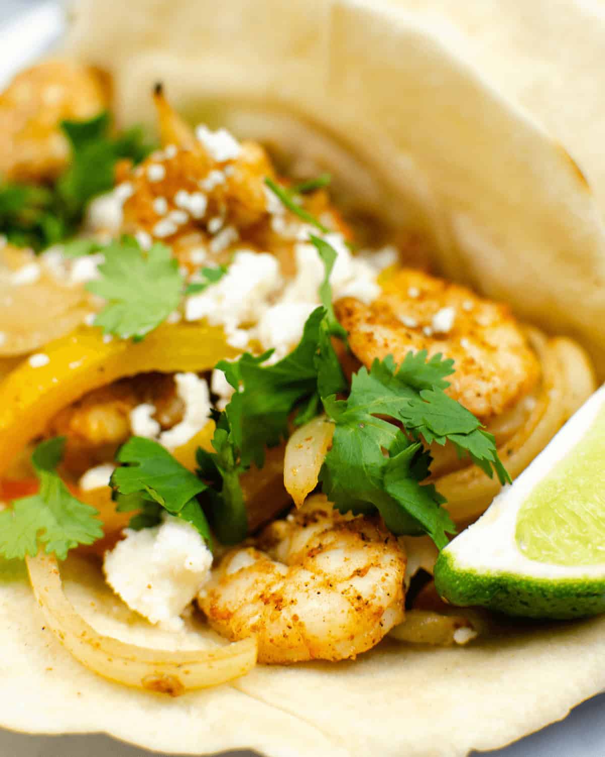 A shrimp fajita taco garnished with cilantro, crumbled cheese, sautéed onions and yellow peppers, served with a lime wedge on a white plate.