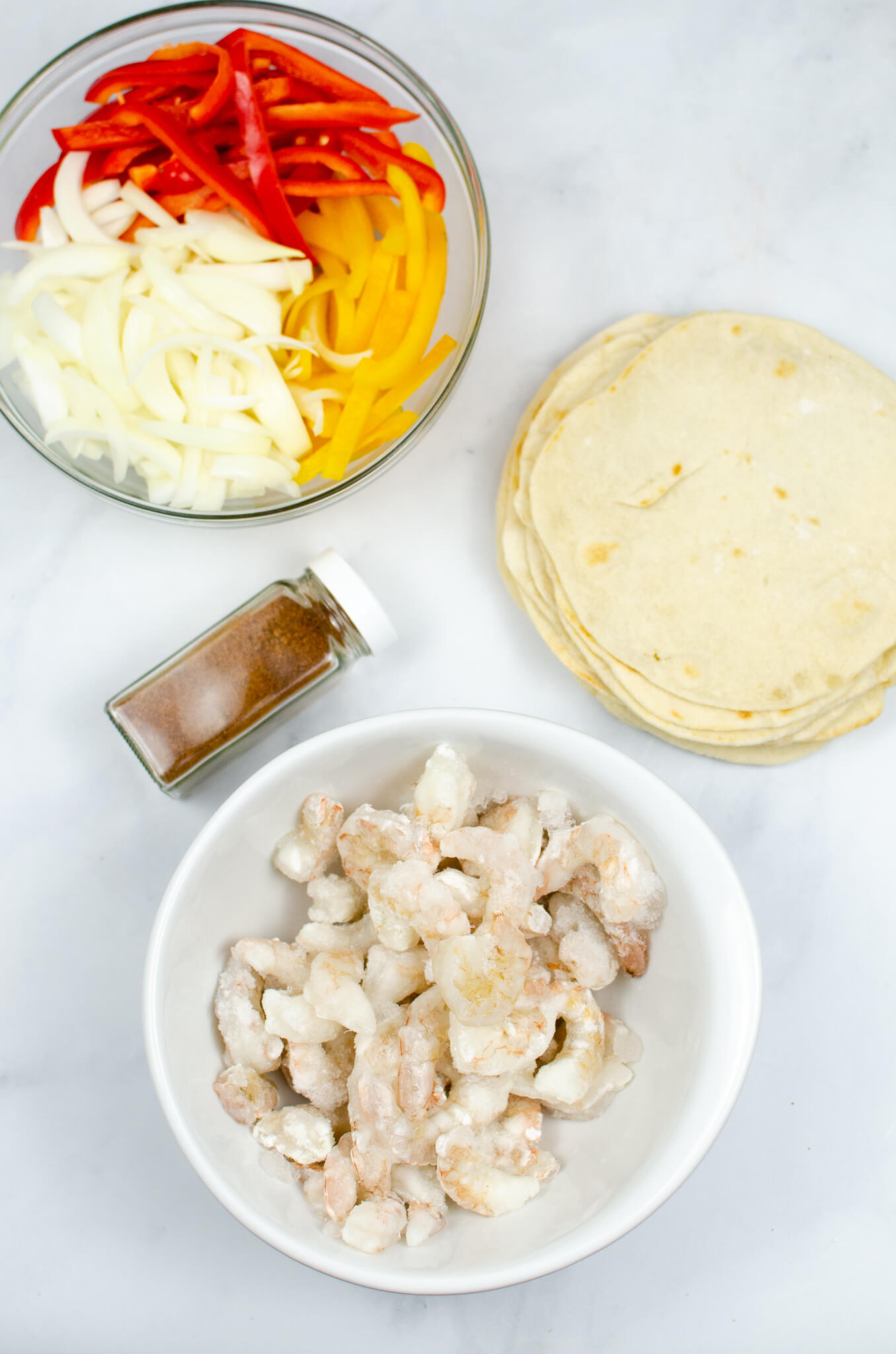 Ingredients for shrimp fajitas including raw shrimp, sliced bell peppers, onions, spices, and tortillas on a baking sheet.