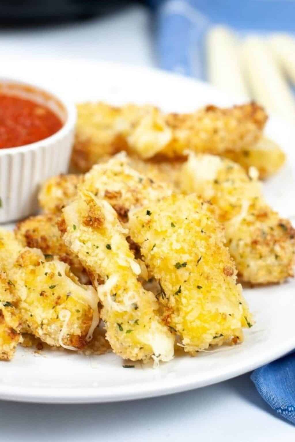 A plate of crispy Air Fryer Mozzarella sticks served with a small bowl of marinara sauce on the side, all arranged on a white plate.