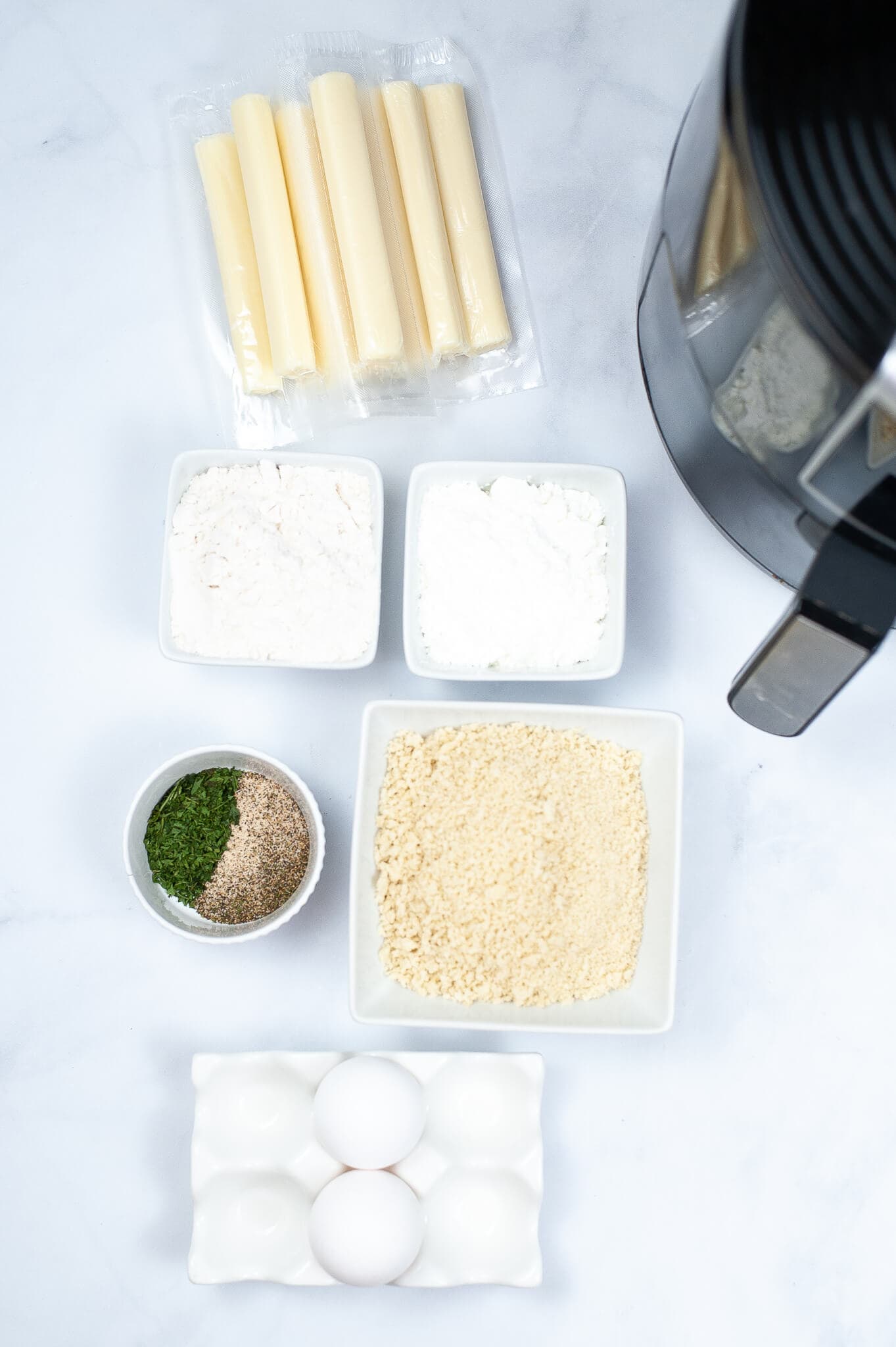 Ingredients for making Air Fryer Mozzarella sticks are laid out on a white surface, including string cheese, flour, cornstarch, bread crumbs, seasonings, eggs, and an air fryer.