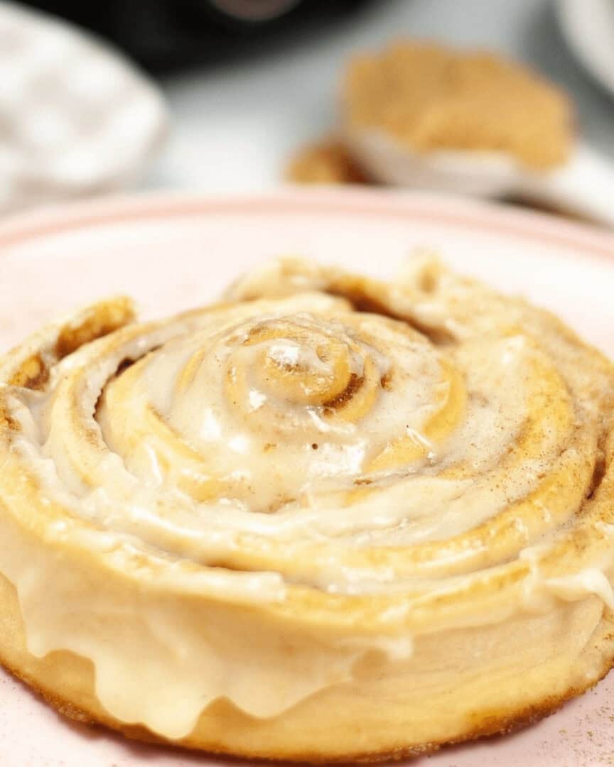 A freshly baked cinnamon roll from an air fryer topped with creamy icing, served on a pink plate, with a blurred background.