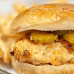 A close-up of a Copycat Chick Fil A Sandwich with pickles on a bun, accompanied by crinkle-cut fries in the background.
