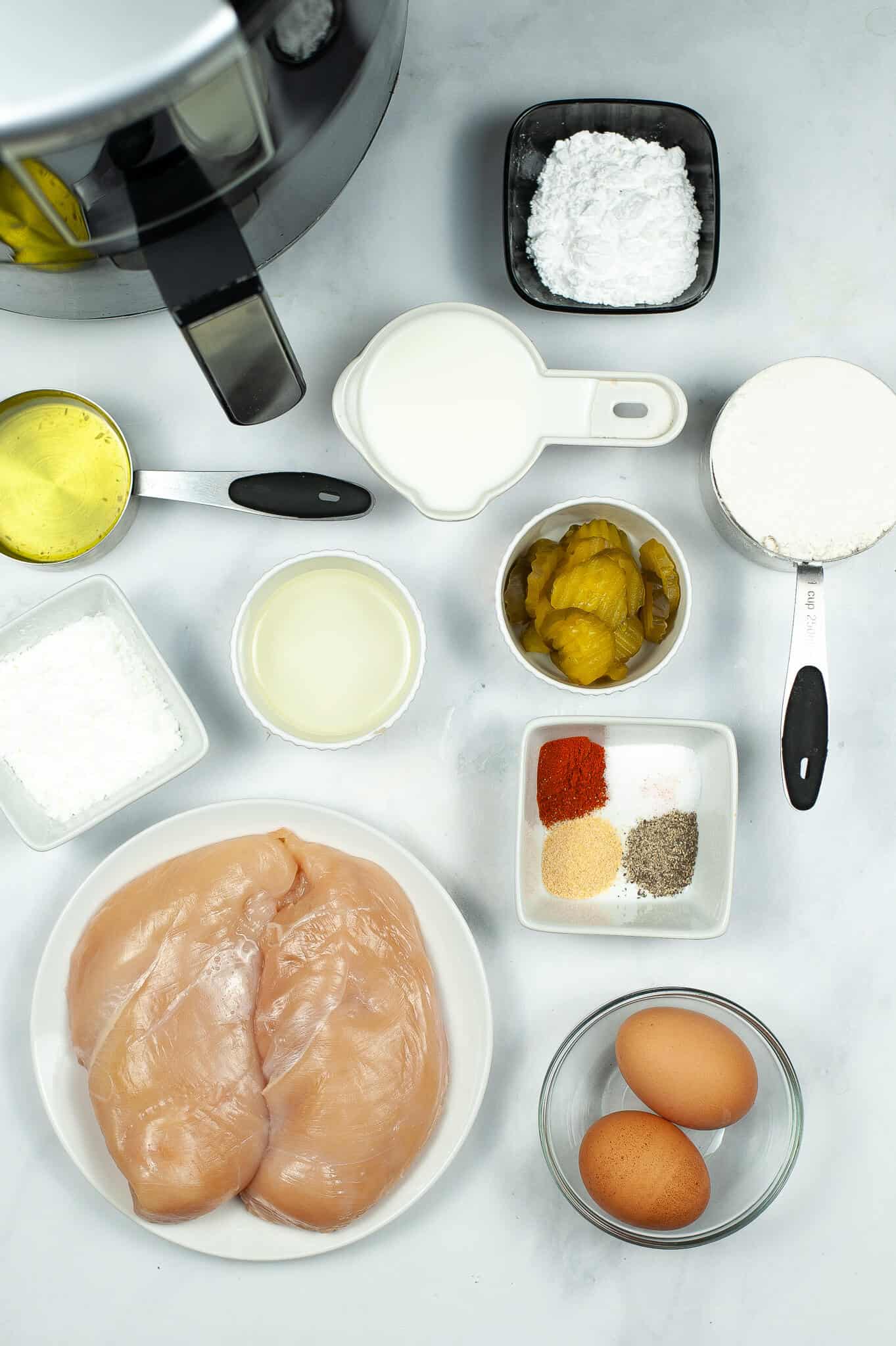 Top-down view of raw chicken breasts, two eggs, various herbs and spices, and other cooking ingredients arranged on a white surface, perfect for making a Copycat Chick Fil A Sandwich. An air fryer is partially visible in the top left corner.