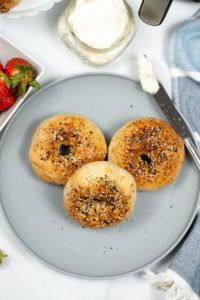 Air Fryer everything bagel on a grey plate with a knife with cream cheese and a hint of strawberries.