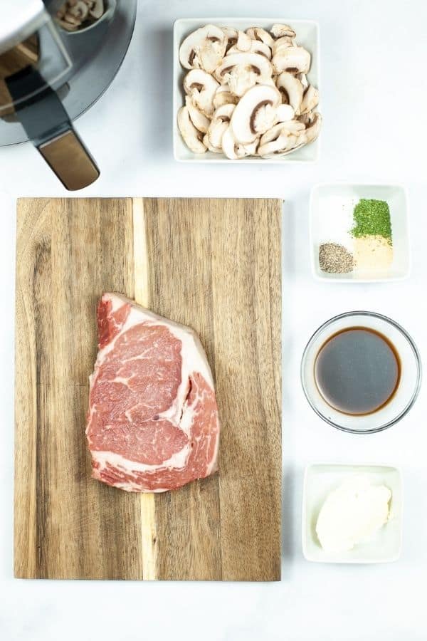 A wooden cutting board with a steak in the middle surrounded by bowls of mushrooms herbs and seasonings.