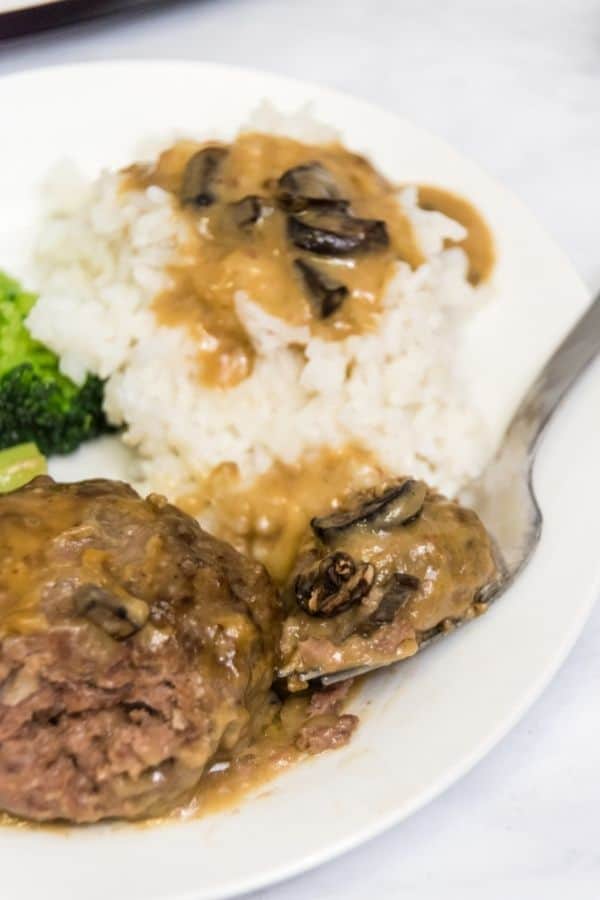 Plate of salisbury steak with a fork on it with broccoli, mashed potatoes and a crock pot filled with salisbury steak in the back.