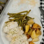 A plate of meat nestled among green beans and roasted potatoes rests beside a black and white checkered napkin. Grated cheese is sprinkled on the table.
