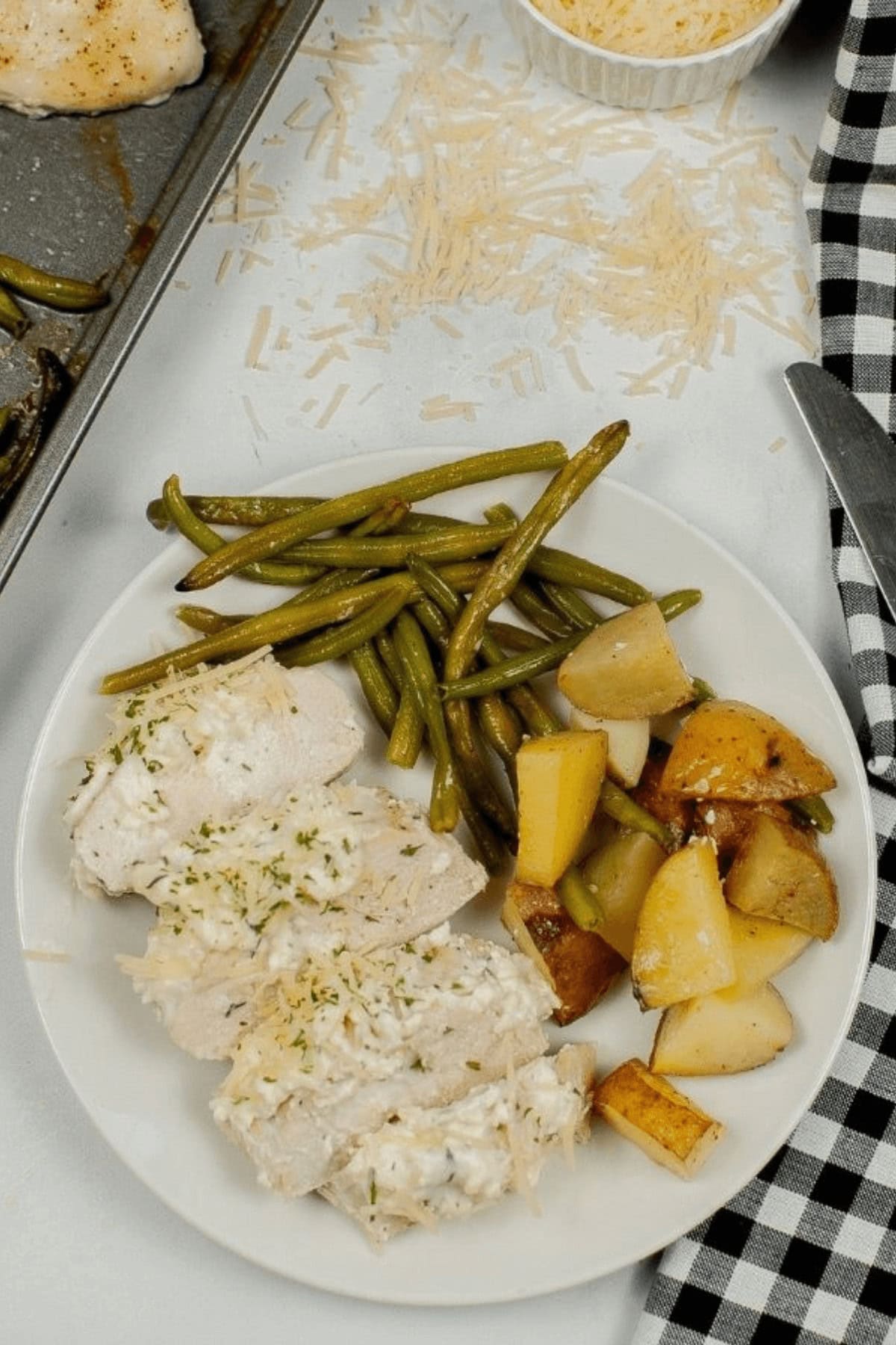A plate of meat nestled among green beans and roasted potatoes rests beside a black and white checkered napkin. Grated cheese is sprinkled on the table.