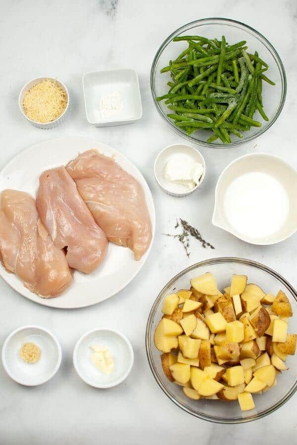 Ingredients for a delicious sheet pan chicken with vegetables: raw chicken breasts, chopped potatoes, green beans, grated cheese, butter, minced garlic, cream, flour, and thyme on a white surface.