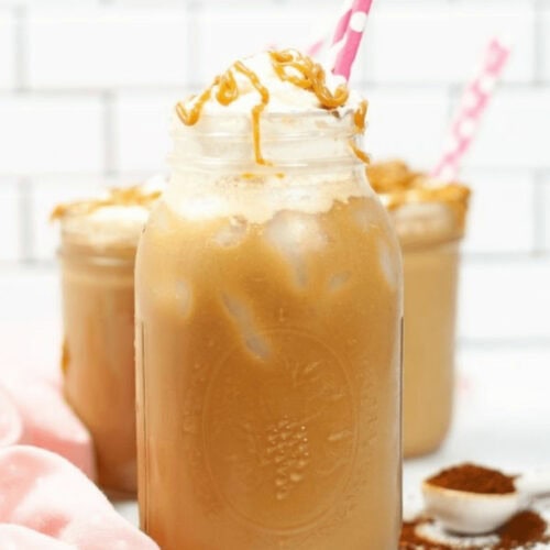 Three Iced Caramel Macchiatos topped with whipped cream and caramel drizzle, served in mason jars with pink and white polka-dot straws.