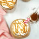 Overhead view of a milkshake topped with whipped cream and caramel drizzle, with pink straws, reminiscent of an Iced Caramel Macchiato. Other milkshakes and a pink cloth are partially visible in the background.
