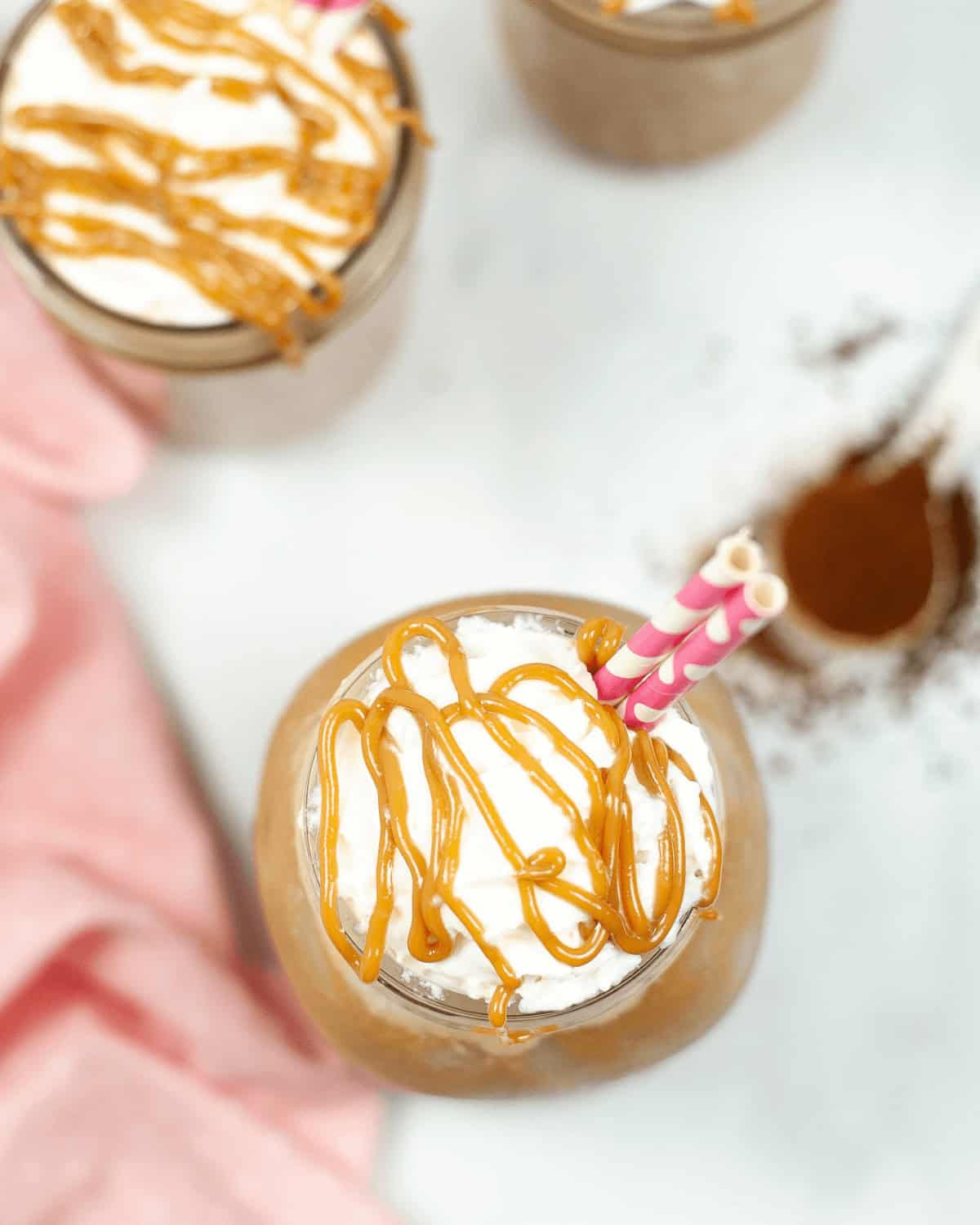 Overhead view of a milkshake topped with whipped cream and caramel drizzle, with pink straws, reminiscent of an Iced Caramel Macchiato. Other milkshakes and a pink cloth are partially visible in the background.