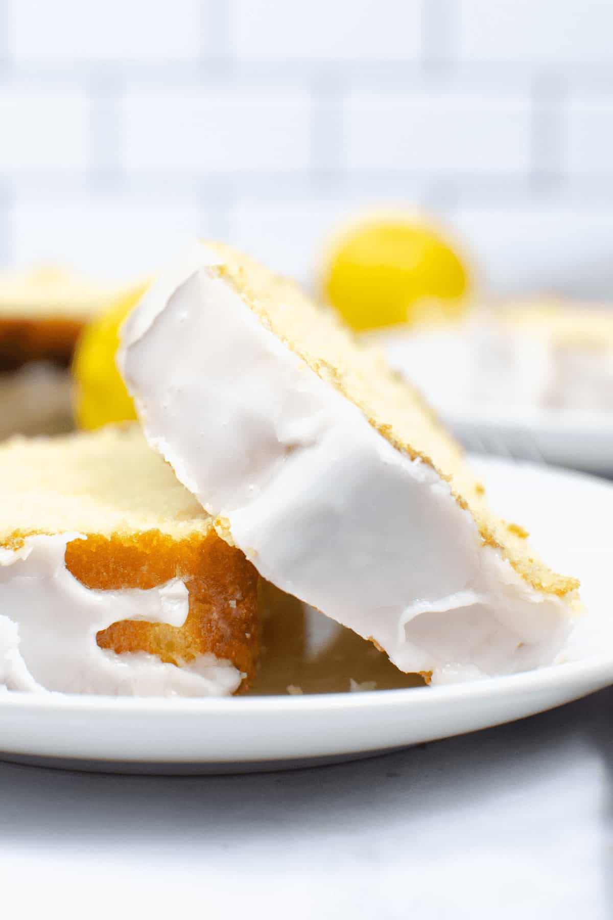 A slice elegantly on a white plate, with a blurred background showcasing fresh lemons and another tempting slice.