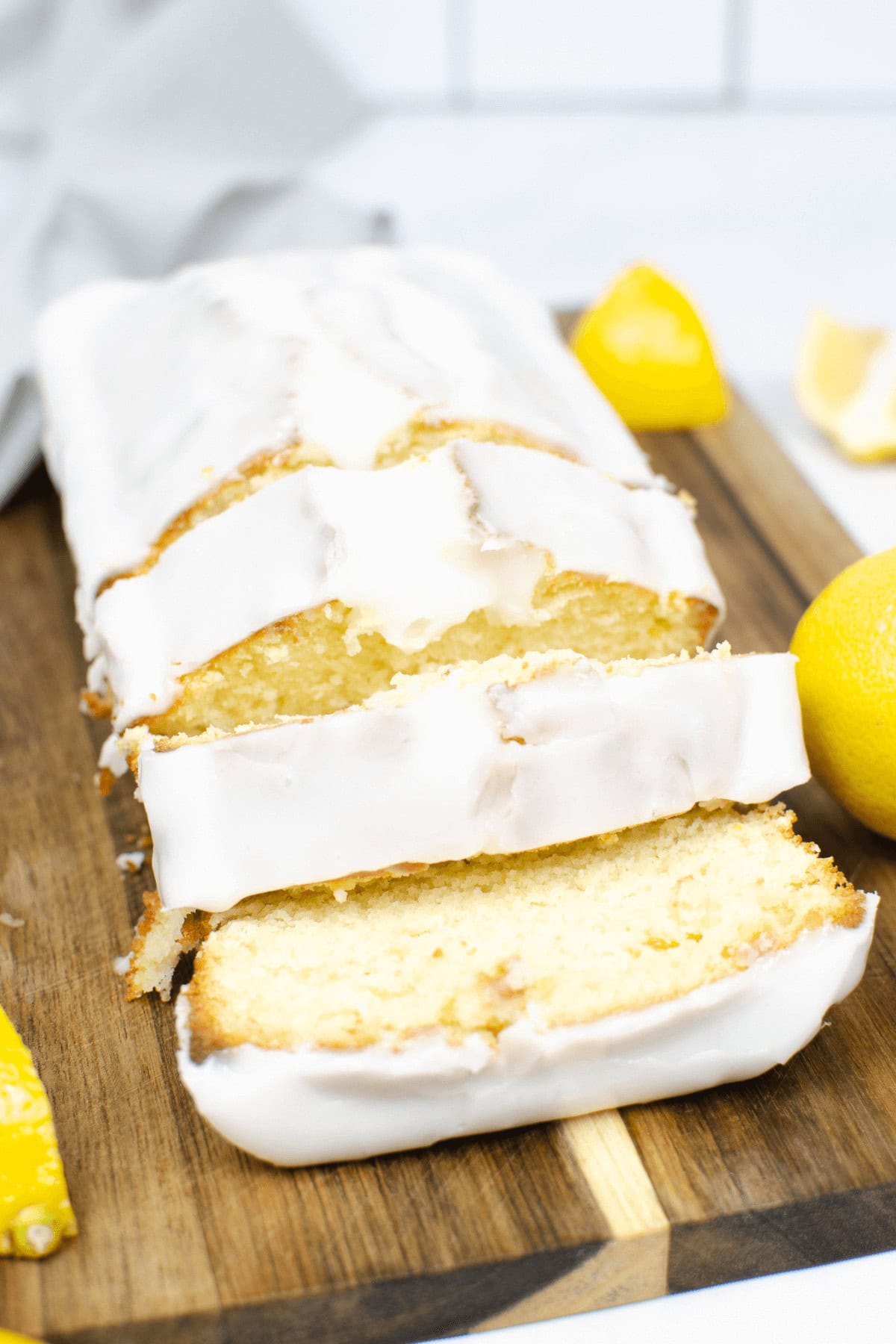 A delightful lemon loaf cake sits on a wooden board, crowned with white icing and surrounded by vibrant lemon halves.