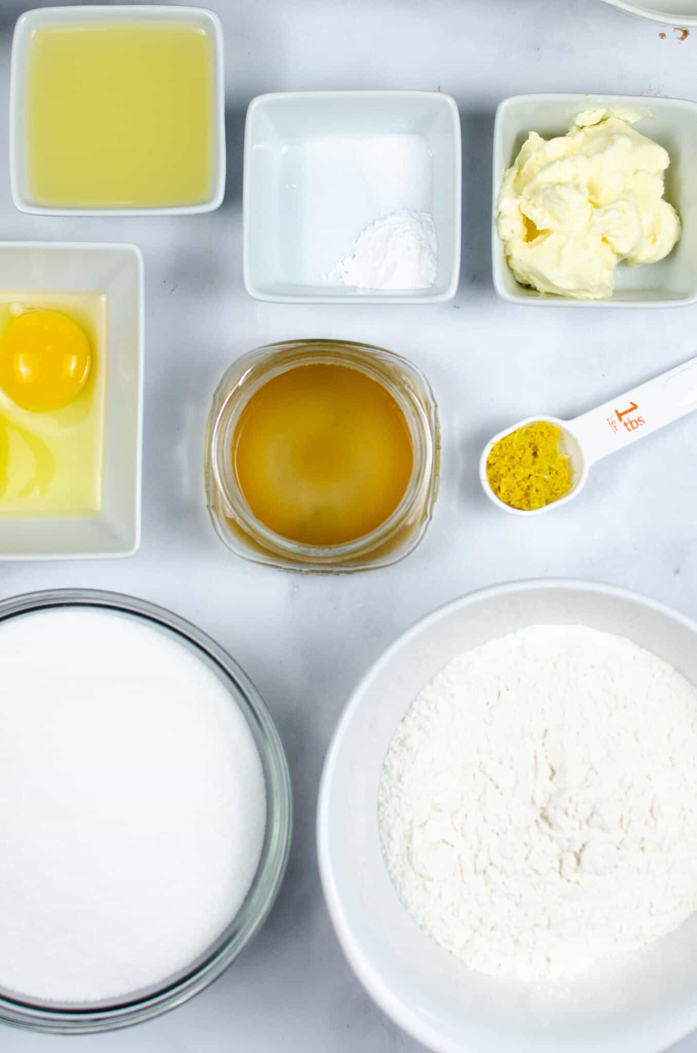 Baking ingredients for a Cake on the countertop include flour, sugar, egg, butter, vanilla extract, lemon zest, baking powder, and lemon juice.