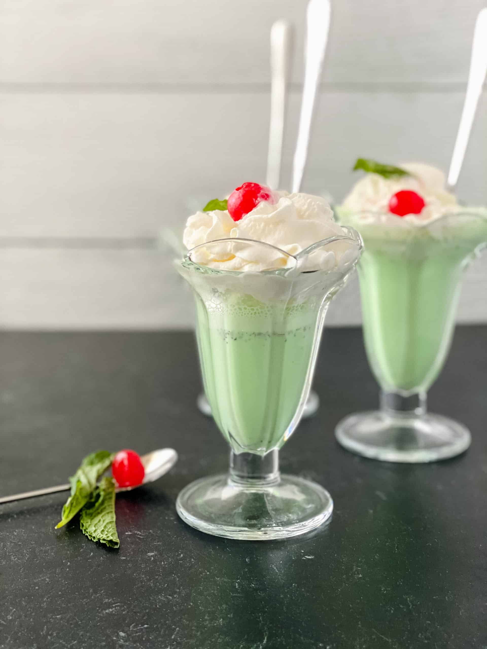 Two shakes on a black counter with a side of mint and a cherry on a spoon