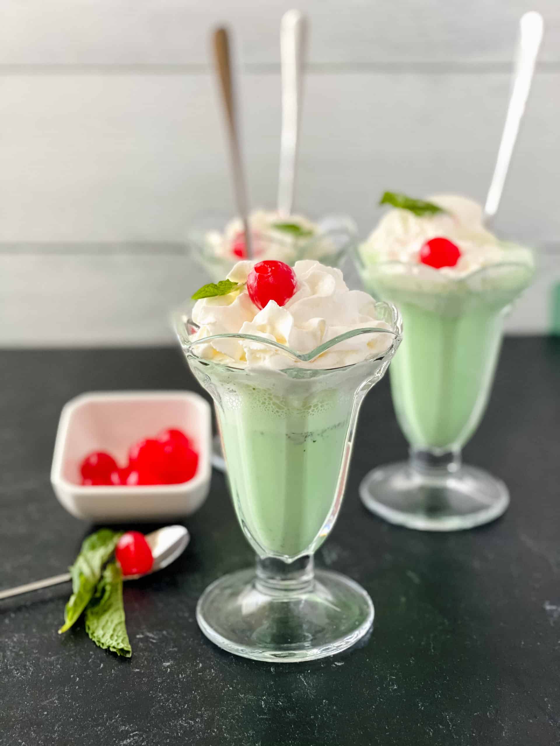 Several Shamrock Shakes with a white dish of cherries on the side