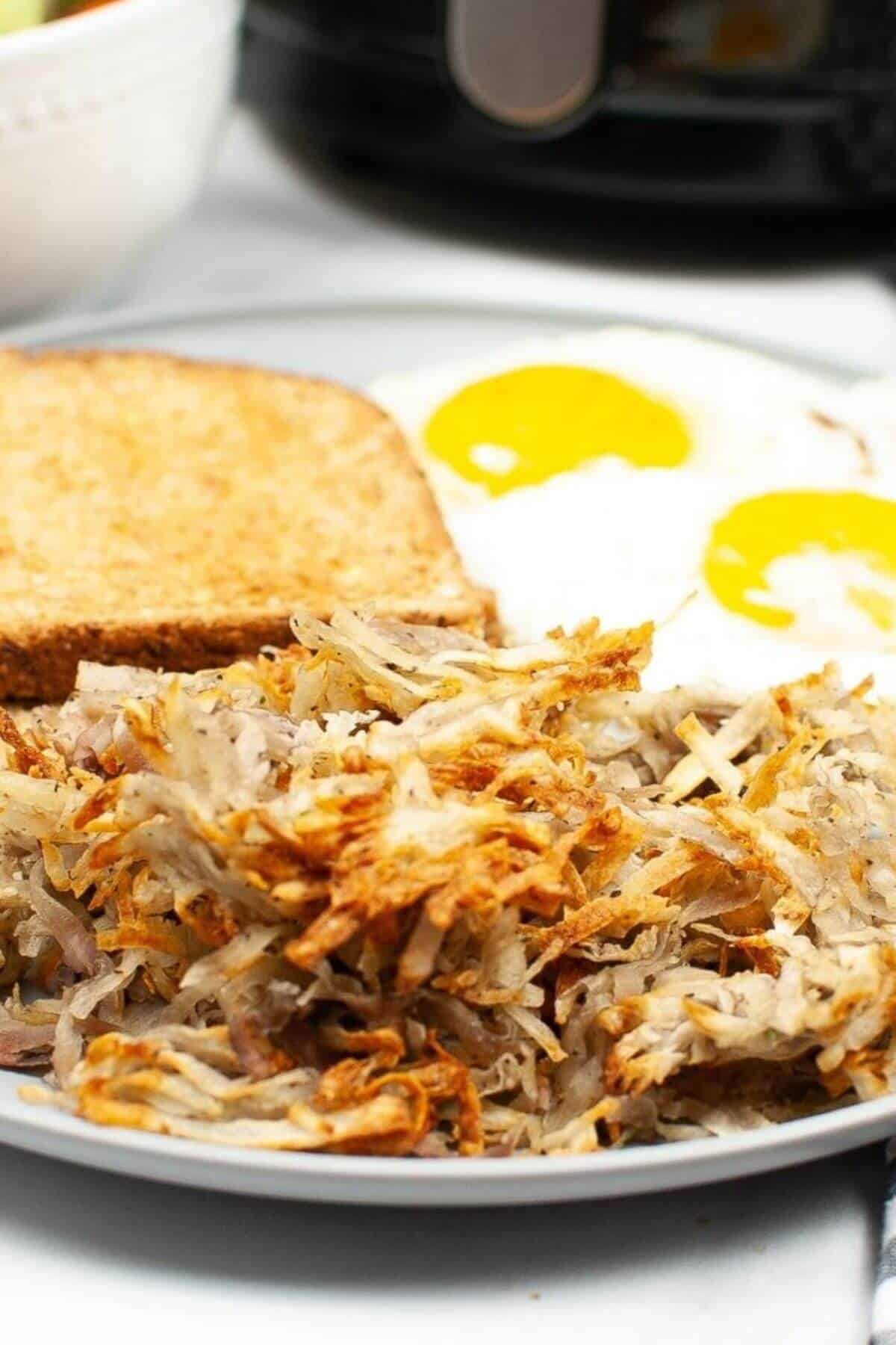 A plate with crispy air fryer hash browns, two sunny-side-up eggs, and a slice of toasted bread.