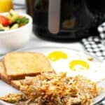 A breakfast plate with potatoes two sunny-side-up eggs, and toast, served alongside a bowl of fruit salad, with an air fryer in the background.