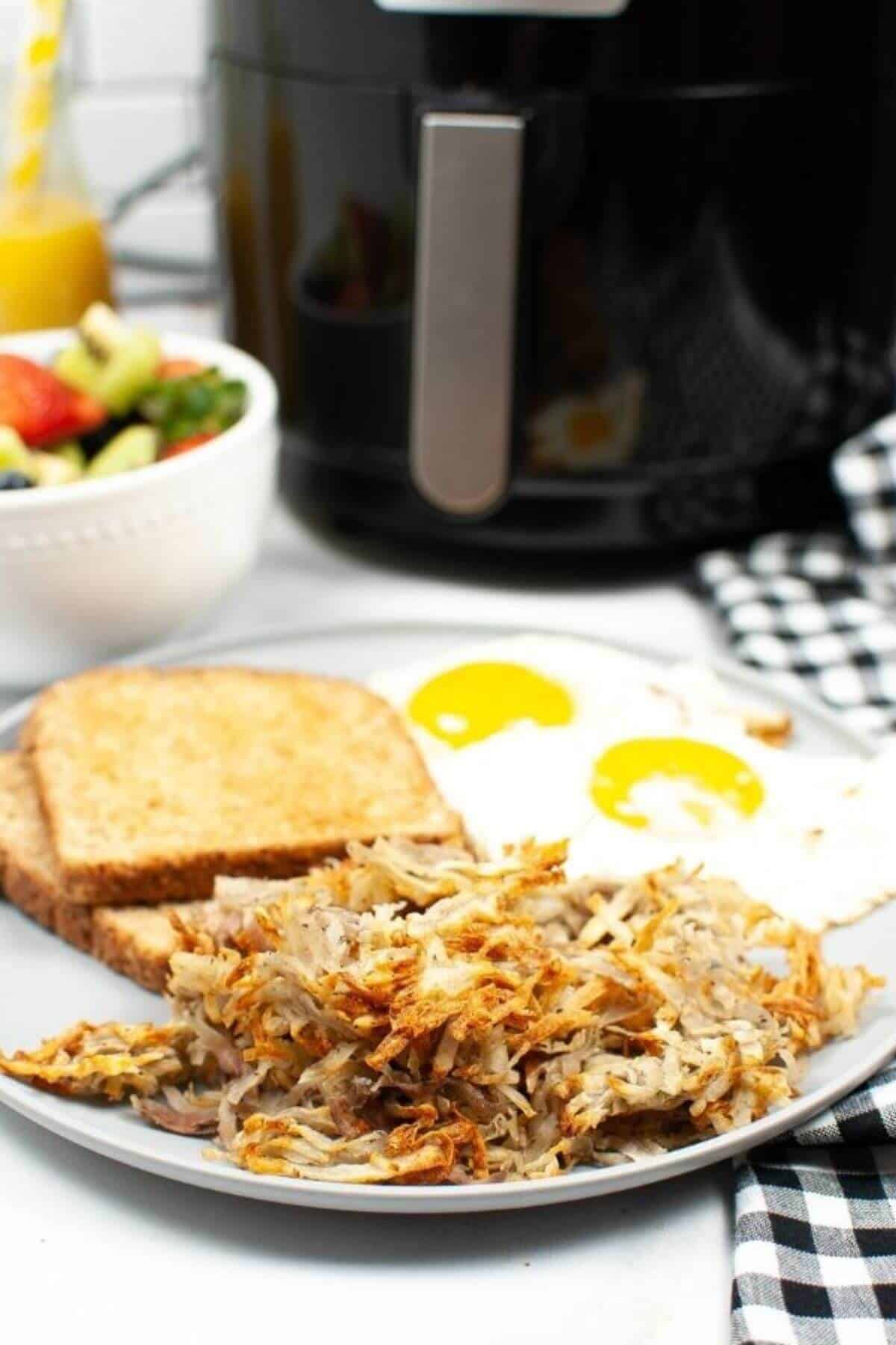 A breakfast plate with potatoes two sunny-side-up eggs, and toast, served alongside a bowl of fruit salad, with an air fryer in the background.