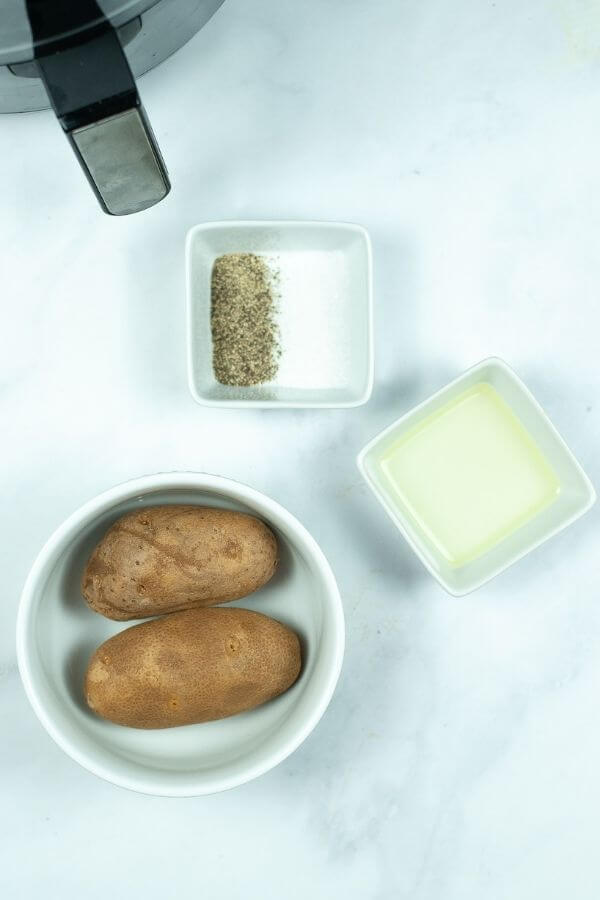 Two whole russet potatoes in a bowl, a small dish of oil, and a small dish with salt and pepper—all the essentials you need are neatly arranged on a white surface.