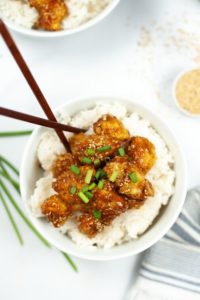 White bowl of rice with sesame chicken on top with chop sticks in the bowl. On a white tablecloth with palm fronds on it