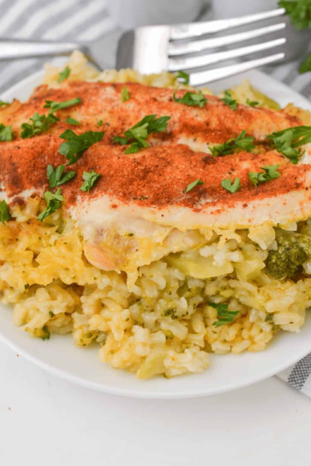 A serving of Cheesy Chicken Broccoli and Rice Casserole topped with paprika and chopped parsley on a white plate, with a fork in the background.